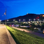 Eine gut beleuchtete Uferpromenade in der Abenddämmerung mit Straßenlaternen, Fahnen und einer Brücke über den Fluss in Trier; im Hintergrund erscheinen Hügel mit beleuchteten Gebäuden unter einem tiefblauen Himmel, die den Charme von SEO Trier widerspiegeln.