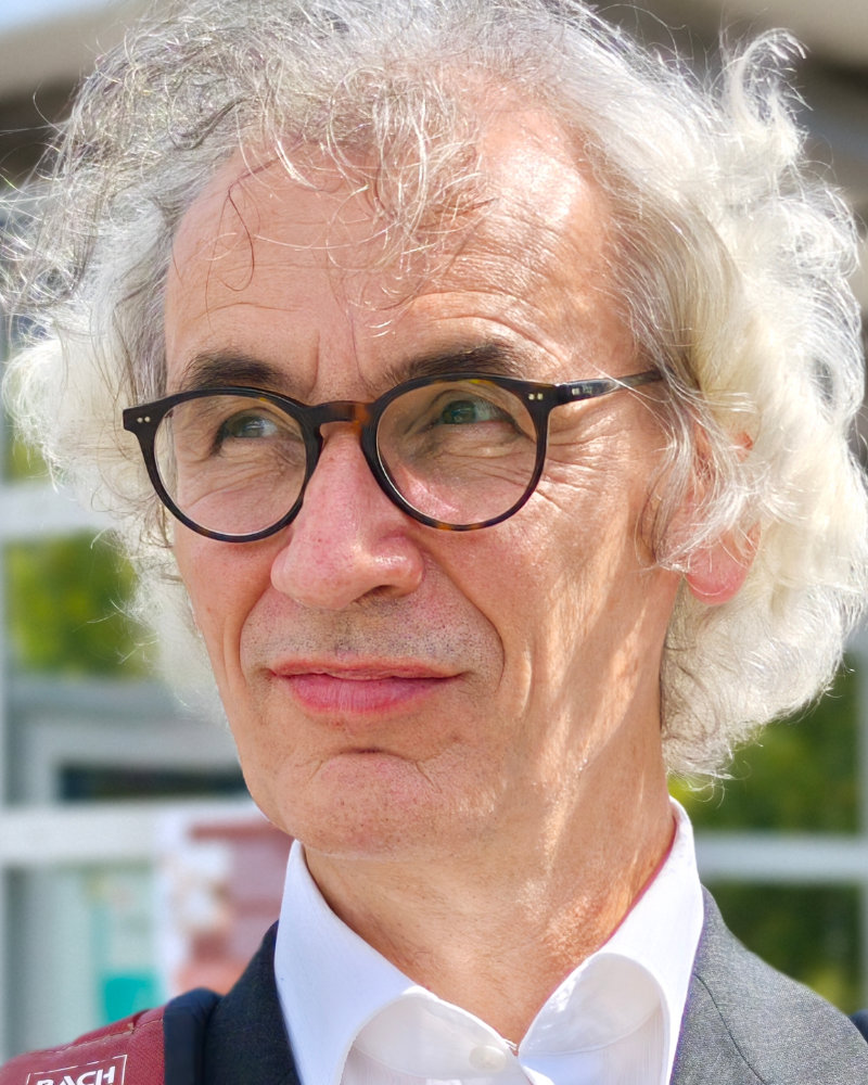Ein älterer Mann mit lockigem, weißem Haar und schwarz umrandeter Brille blickt nachdenklich zur Seite. Er trägt ein weißes Hemd und eine dunkle Jacke und steht im Freien im Sonnenlicht - vielleicht denkt er über sein nächstes Projekt bei der Webagentur Trier nach.